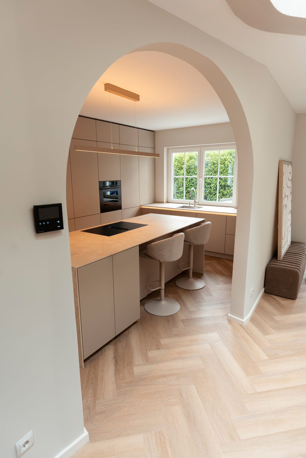 Blick durch den weißen Rundbogen auf die sandbeige Inselküche mit Barhockern und Fensterzeile