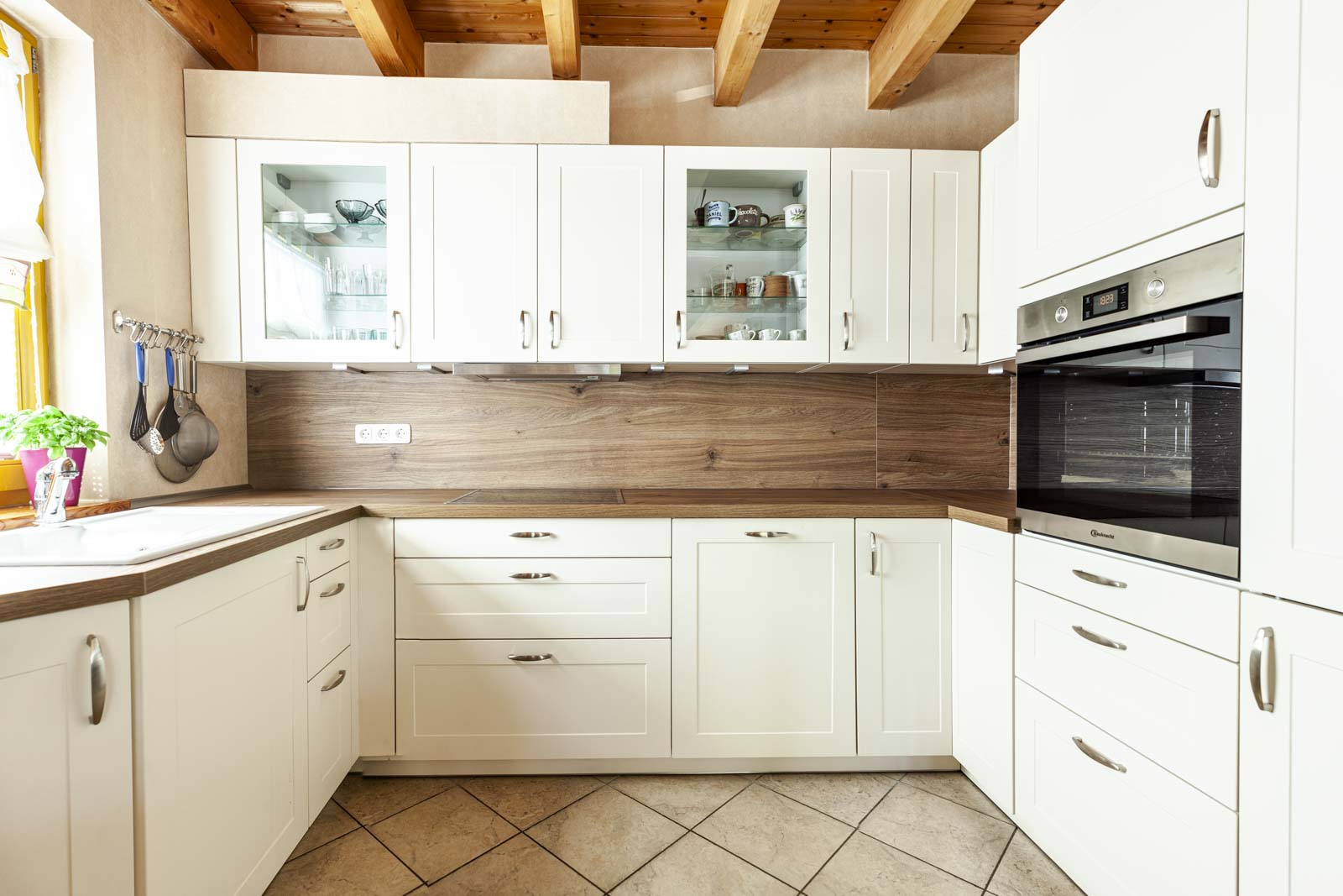 Helle U-Landhausküche mit Rahmenfronten in Magnolie, Nischenrückwand in Eiche Marone und Holzbalkendecke