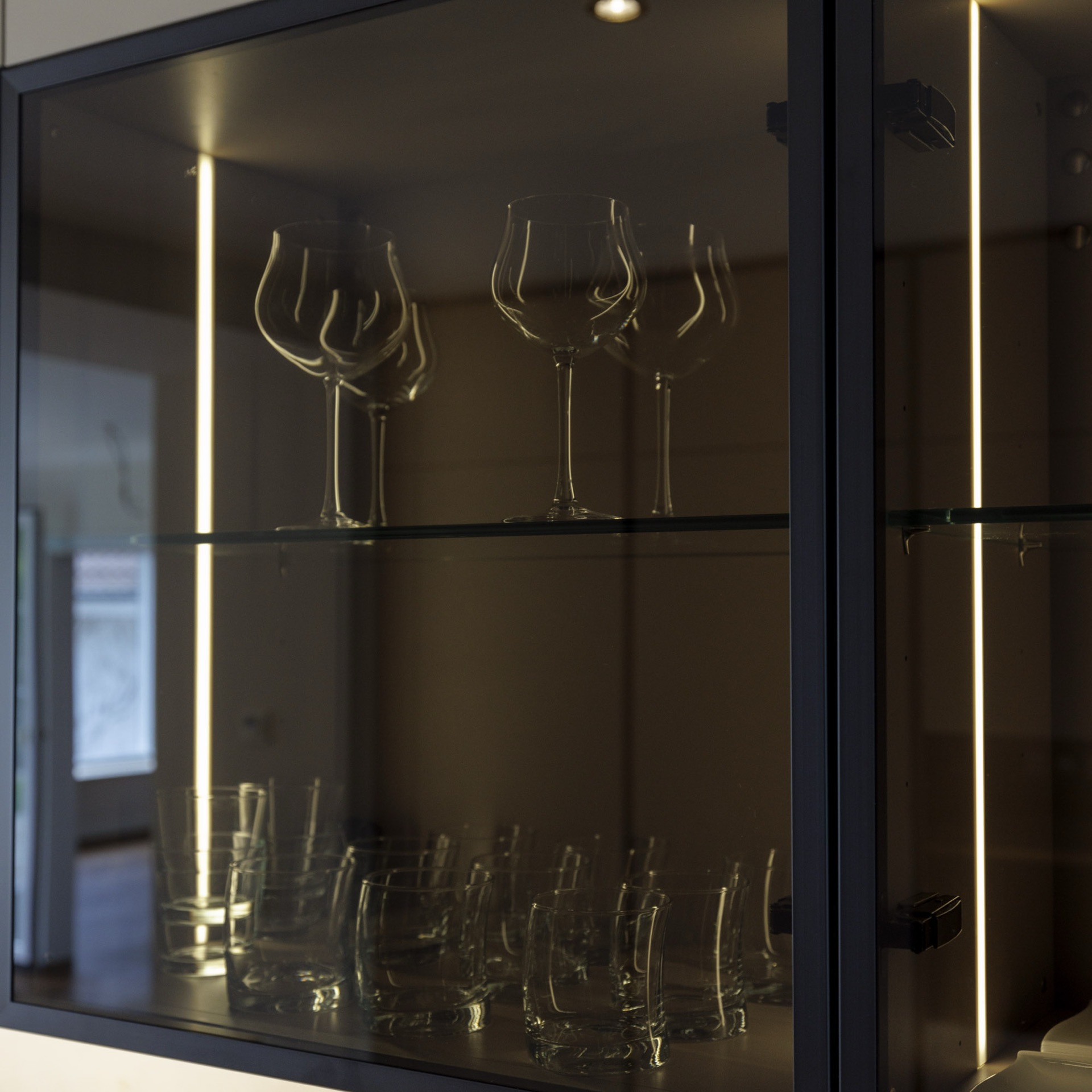 Glashängeschrank mit schwarzem Rahmen und LED-Innenbeleuchtung, davor Gläser auf Glasböden